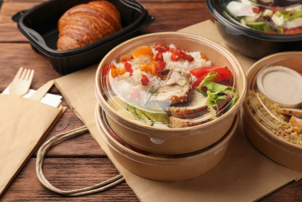 Tasty food in containers with knife and fork on a wooden table, close-up.